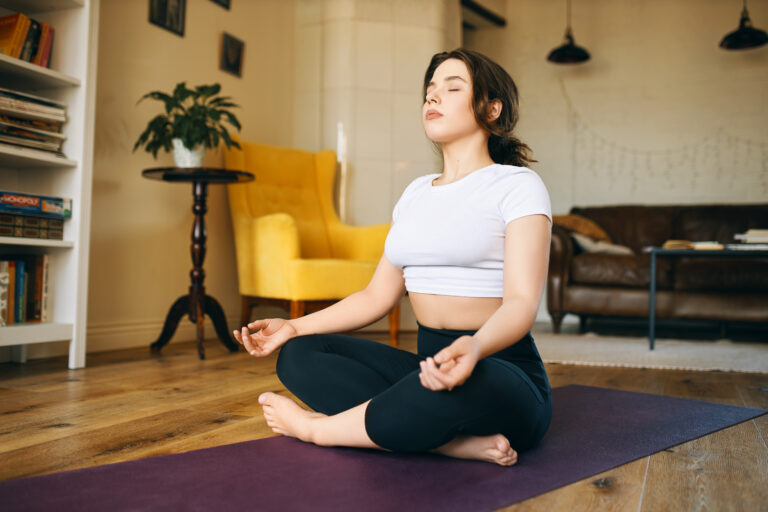 Pretty with chubby cheeks sitting on yoga mat with legs folded, closing eyes, taking deep breath, practicing meditation, searching for inner peace and harmony